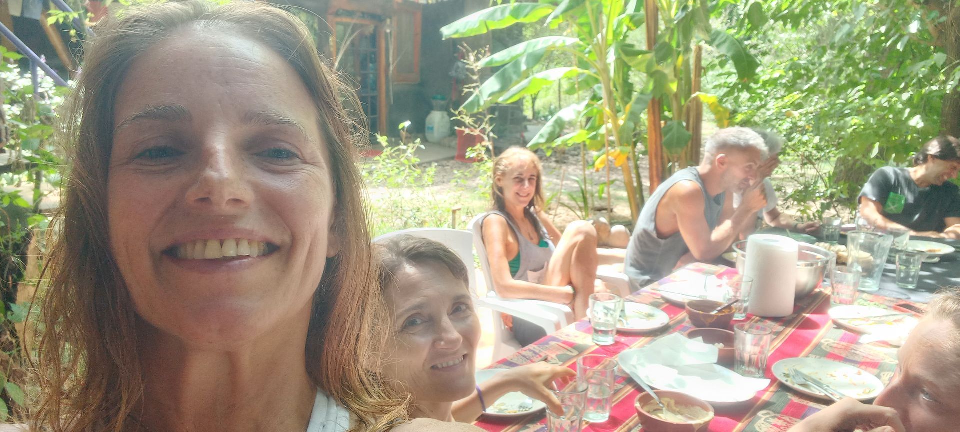La gente se reunió al aire libre, sonriendo y comiendo en una mesa con comida, rodeada de exuberante vegetación.