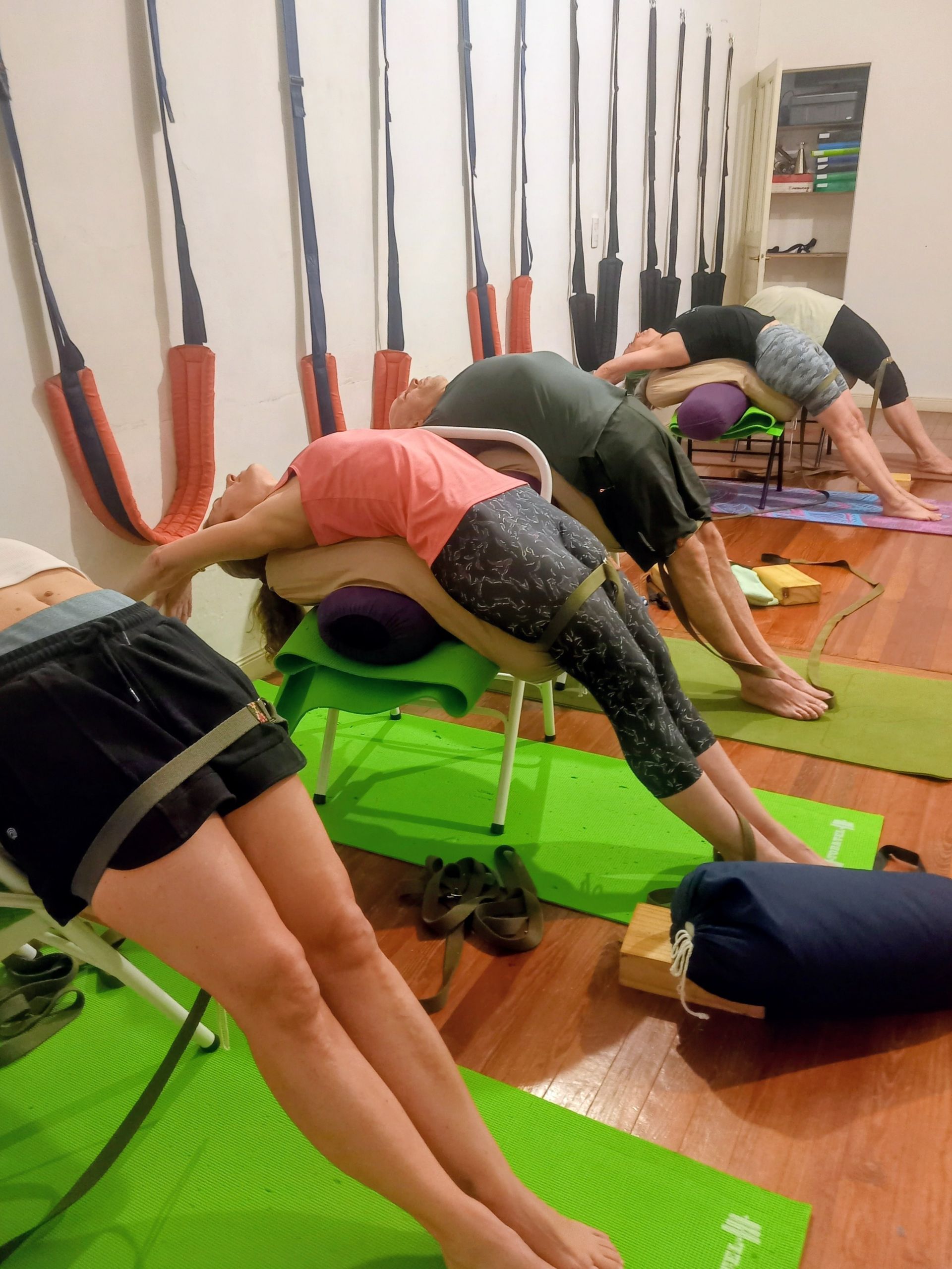 Personas en una clase de yoga arqueando la espalda sobre sillas, con colchonetas, cojines y correas de pared en un estudio.