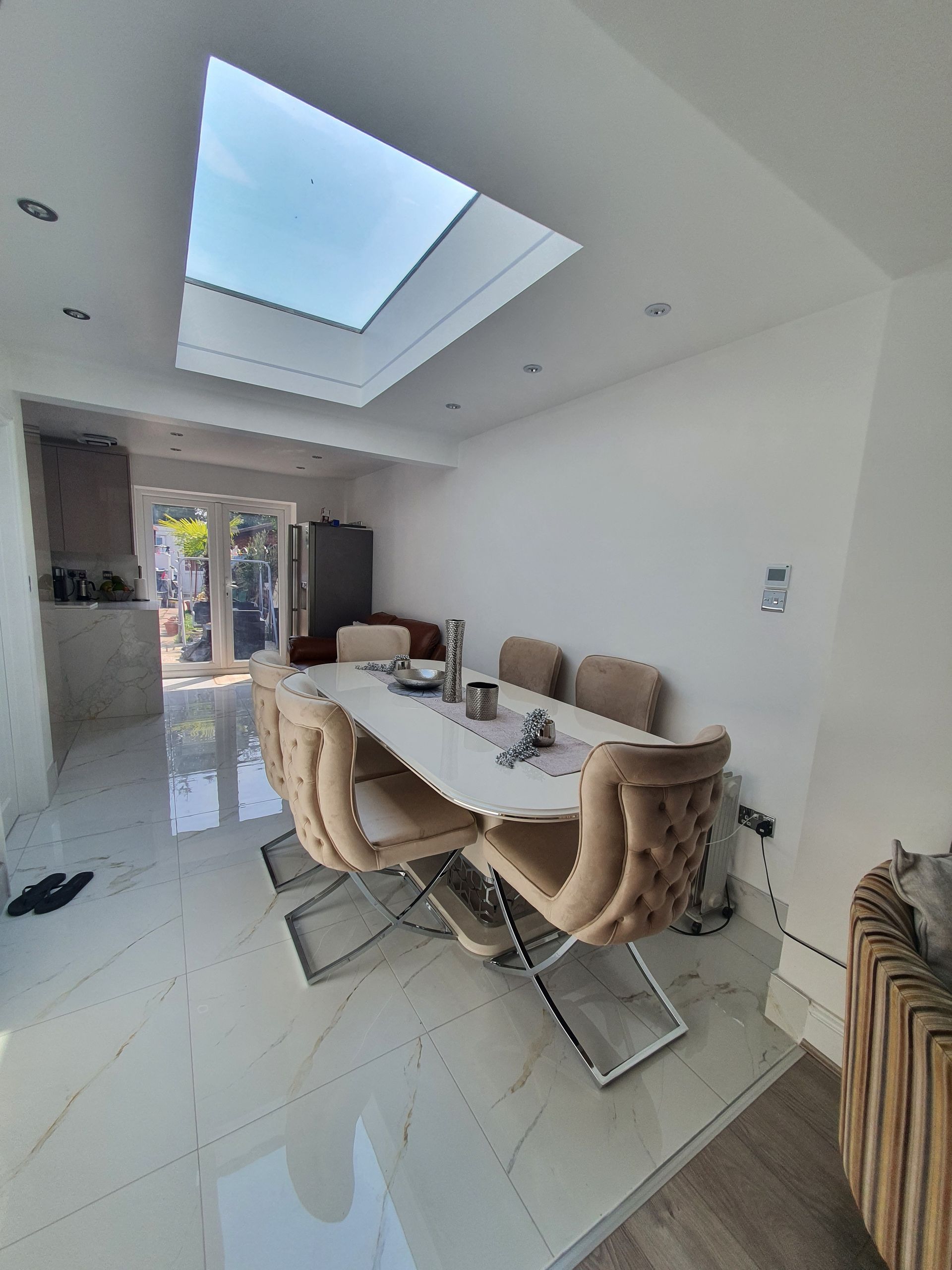 A dining room with white tiled floors, a long white table, and six beige tufted chairs under a large ceiling skylight.