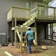 A person in a light blue shirt stands on a wooden ladder under a partially constructed deck stairway at the back of a house.
