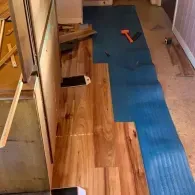Wood-look laminate planks being installed on a blue foam underlayment in a room under construction.