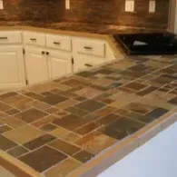 A kitchen counter covered in brown, tan, and grey square stone tiles, next to white cabinets and a dark backsplash.