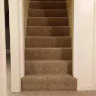 A straight flight of stairs carpeted in a neutral tan color, positioned between two plain, off-white walls.