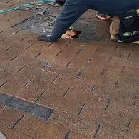 A person in a dark jacket and gloves kneels on a brown shingled roof, repairing a damaged section of asphalt shingles.