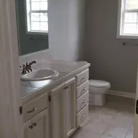 A modern bathroom featuring a white vanity with a silver faucet, a white sink, and a toilet against a light grey wall.