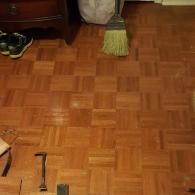 A medium-angle shot of a parquet wood floor with a broom, hammer, and shoes scattered around the room.