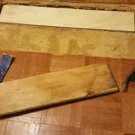 A parquet floor repair featuring a removed section with exposed subflooring, a replacement wood plank, and a pry bar.