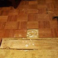 Parquet wood floor tiles with one tile missing, exposing the glue and wooden subfloor underneath.