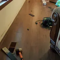 A hammer and a floor vent sit on a dark-toned wood floor in a room under construction, with tools scattered nearby.