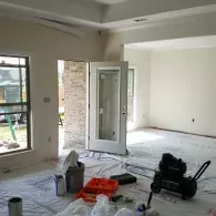 An interior room under construction with plastic sheeting on the floor, a door leading to brickwork, and an air compressor.