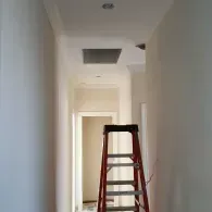 A red stepladder stands in a narrow, white-painted hallway leading to an open doorway.