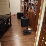 A wood-floored room featuring a wooden media cabinet filled with movies, two black subwoofers, and some scattered items.