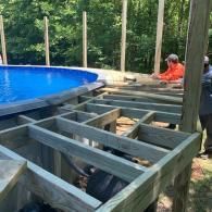 A person working on the wooden framing for a deck surrounding an above-ground swimming pool in a wooded setting.