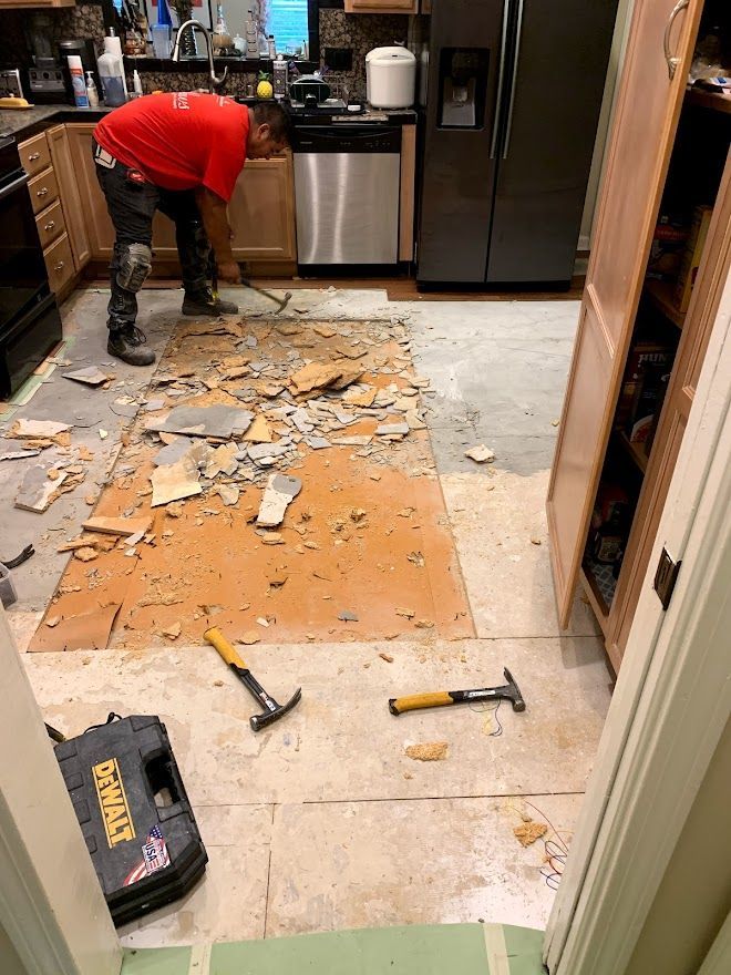 A worker in a red shirt uses a hammer to remove floor tiles in a kitchen, with tools and debris scattered on the floor.