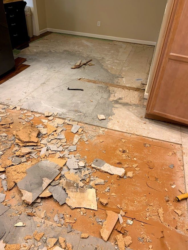 A room undergoing a floor renovation with old tile debris scattered across a brown subfloor and exposed concrete.