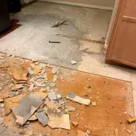 A room floor in the process of demolition, showing exposed concrete, scattered tiles, and debris.