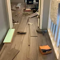 Wood-look flooring planks are partially installed on a hallway floor with tools, packaging, and remnants scattered around.