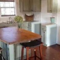 A kitchen island with a wooden countertop and a stool sits in front of light-green cabinets and a window.