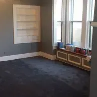 A gray room with a recessed white wall shelf, a dark carpet, and a window featuring a radiator cover lined with books.