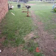 A gas-powered post hole digger stands upright in a lawn with a row of freshly dug holes stretching into the yard.