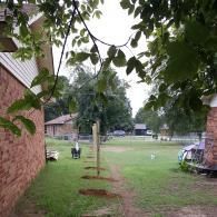 A residential backyard view shows a line of wooden posts set in the ground for a new fence, framed by brick walls.