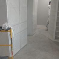 A person in white work clothes paints a wall in a room featuring white built-in cabinets and a yellow metal scaffold.