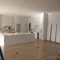 A worker paints the walls and island of a large kitchen with white primer, surrounded by newly installed cabinetry.