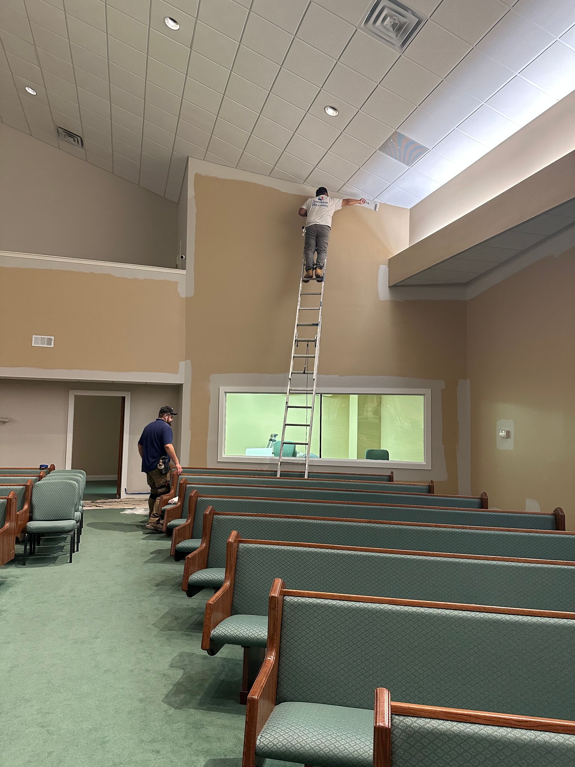 Two workers renovate a church sanctuary, with one on a tall ladder painting the high wall near the ceiling.