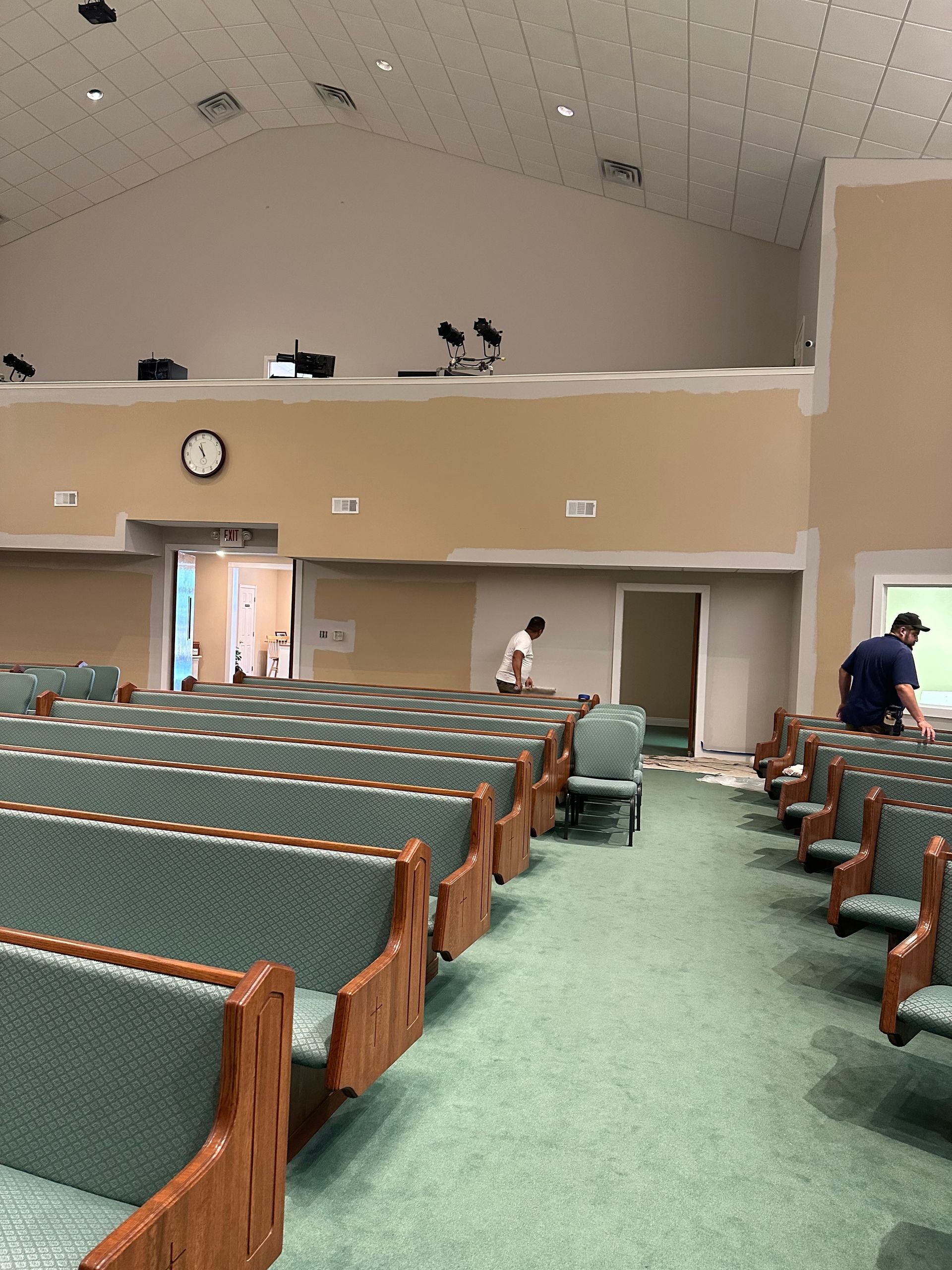 An indoor view of a church sanctuary with rows of green-cushioned pews, beige walls under renovation, and two people working.