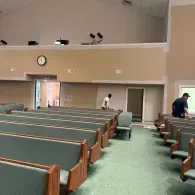 An interior view of a church sanctuary with rows of green-cushioned pews, beige walls, and two people working inside.