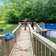 Two people standing on a wooden deck ramp leading to an above-ground swimming pool in a grassy, wooded backyard.