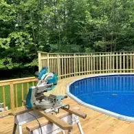 A miter saw rests on a wooden workbench on a deck next to an above-ground pool, with lush green trees in the background.