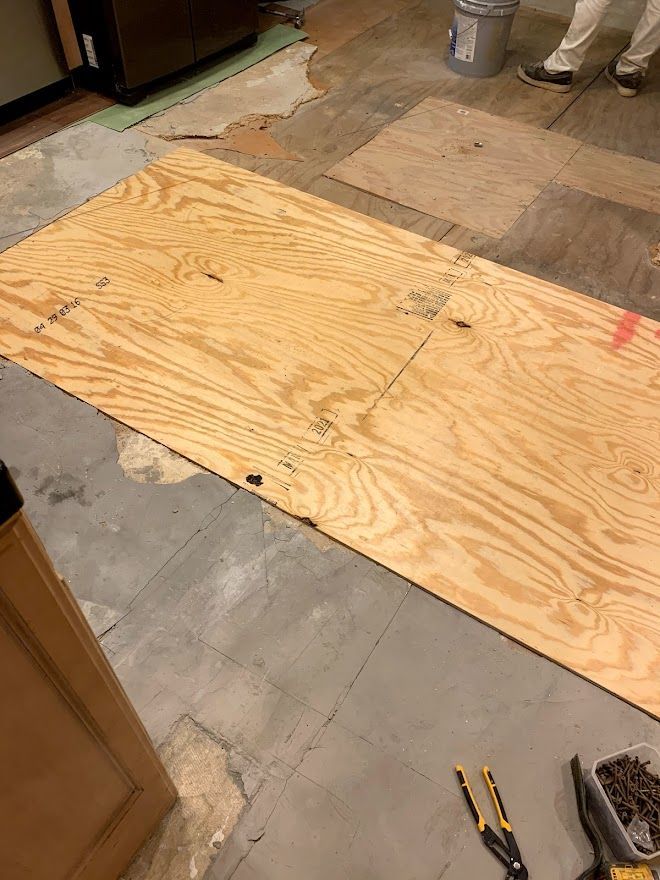 A large sheet of plywood sits on a kitchen floor partially covered with existing vinyl tiles during a renovation project.