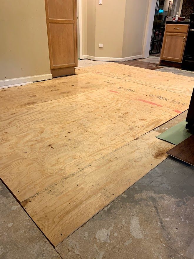 Plywood subflooring installed over a concrete base in a kitchen area with cabinets visible in the background.