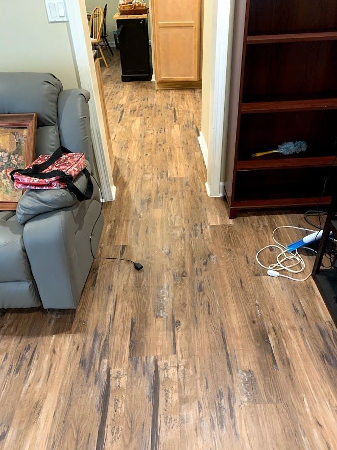 Wood-look vinyl flooring spans a living room and hallway, featuring a grey sofa, a wooden bookcase, and some loose cords.