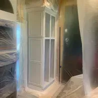 A freshly painted white cabinet stands in a room covered in protective plastic sheeting during home renovations.