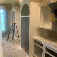 An interior room under construction features white built-in cabinets, an arched alcove, and a stepladder on a drop cloth.