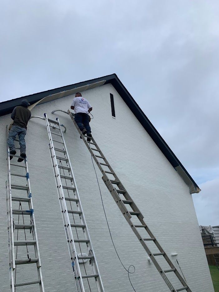 Two workers on extension ladders repair the white siding and trim near the roofline of a two-story building.