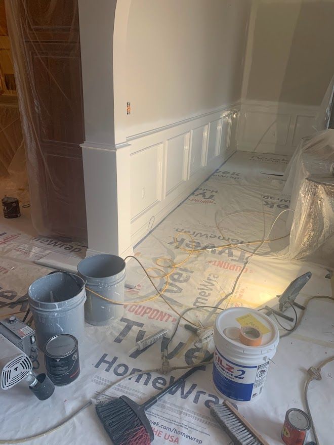 A construction site with white wainscoting on a wall, surrounded by paint buckets, tools, and floor covering.