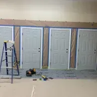 Four light gray doors lined up against a wall, with masking paper taped around them and a step ladder in the foreground.
