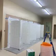 A row of white interior doors stands in an unfinished room with protective plastic sheeting and a ladder nearby.