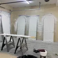 Four white arched doors stand in a garage, lined up for painting against a plastic-covered wall with a workbench nearby.