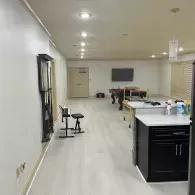 A minimalist open-plan room with light wood floors, a black piano stool, a pool table in the distance, and dark cabinets.