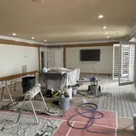 A room undergoing renovation with walls taped off, plastic-covered furniture, painting equipment, and blue hoses on the floor.