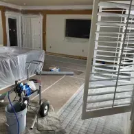A room under renovation with a paint sprayer, a bucket, covered furniture, and white plantation shutters.