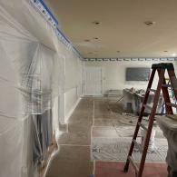 A room under renovation with walls and windows covered in plastic sheeting, a ladder in the foreground, and taped trim.