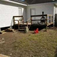 A white house with a raised wooden deck, outdoor fireplace, stairs, and a red plastic chair on a grassy yard.