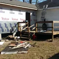 A house under exterior renovation with exposed house wrap, a wooden deck, stairs, and construction debris on the ground.