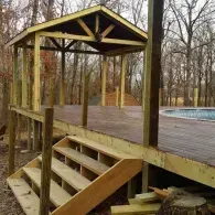 A wooden deck under construction with stairs in the foreground, a roofed gazebo area, and an above-ground pool.