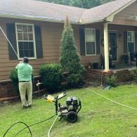 A person in a green shirt uses a pressure washer to clean the exterior of a house, standing near a pressure washer machine.